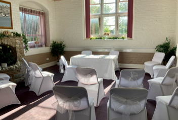 The Clun Room set up for a ceremony as viewed from the back of the room. There is a registry table with two chairs behind it. Facing this are curved rows of chairs with an aisle down the centre. The chairs are decorated with white chair covers and white sashes tied in bows. The room is decorated with potted bay trees and flower arrangements on the fireplace mantlepiece and in the hearth. 