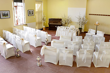 The Severn Wye Room set up for a ceremony as viewed from the back of the room. There is a registry table with two chairs to the side. Facing this are curved rows of chairs with an aisle down the centre. The chairs are decorated with white chair covers and white sashes tied in bows. The aisle is lined with lanterns on the floor.The room is decorated with white bloosoming trees in pots and flowers along the front of the registry table. There is a piano behind and to the left of the registry table.