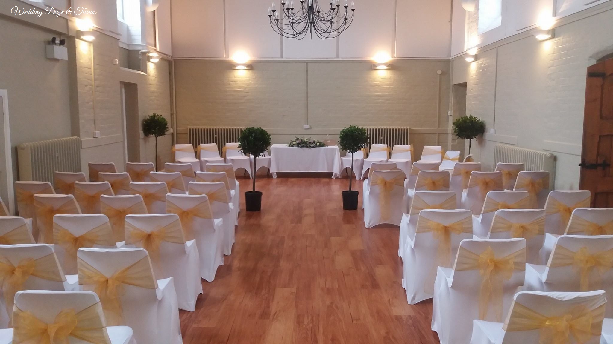 The Hall set up for a ceremony as viewed from the back of the room. There is a registry table with four chairs on either side. Facing this are rows of chairs with an aisle down the centre. The chairs are decorated with white chair covers and gold sashes tied in bows. The room is decorated with potted bay balls and a flower arrangement on the registry table.