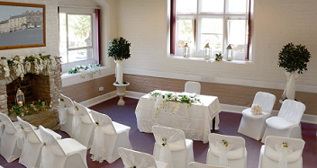 The Clun Room set up for a ceremony as viewed from the back of the room. There is a registry table with two chairs behind it. Facing this are curved rows of chairs with an aisle down the centre. The chairs are decorated with white chair covers and white sashes tied in bows. The room is decorated with potted bay trees and flower arrangements on the fireplace mantlepiece and in the hearth. 